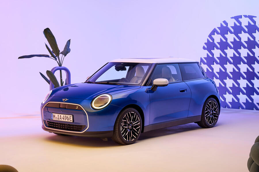 A blue 2024 Mini Cooper SE in a studio with orange and purple lighting, parked in front of a potted plant. Front quarter static