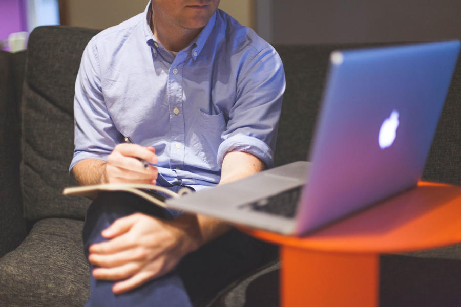 Une personne en chemise bleue prend des notes manuscrites tout en regardant un ordinateur portable Macbook
