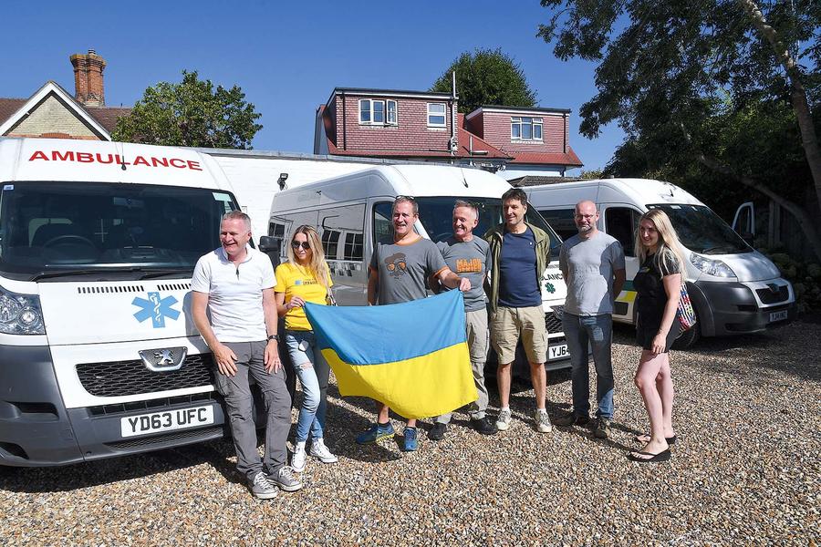 A group of people hold up a Ukranian flag in front of three parked Peugeot ambulances they will donate to Ukraine